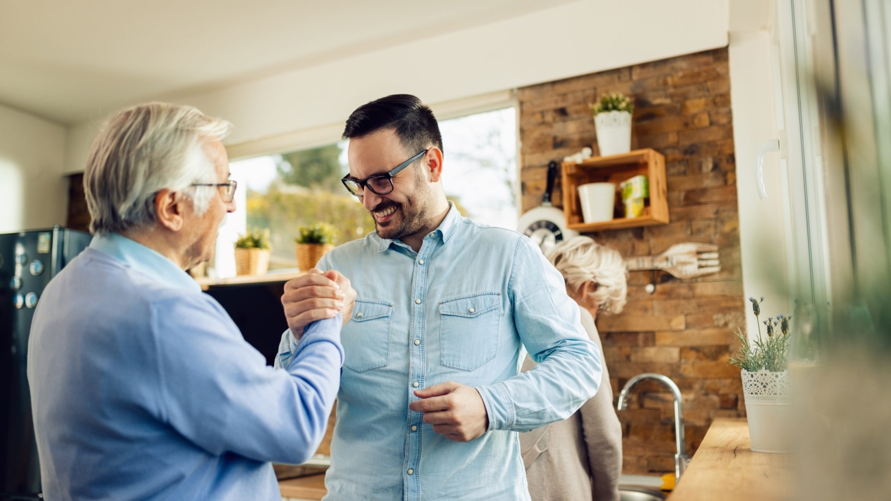 Happy adult son and his mature father holding hands while greeting each other in the kitchen.