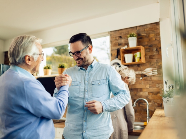 Happy adult son and his mature father holding hands while greeting each other in the kitchen.